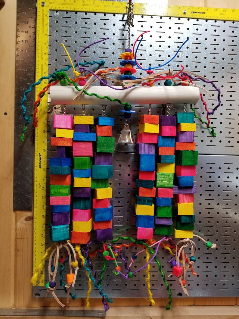May include: A colorful wooden bird toy with multiple hanging blocks, beads, and strings. The toy is suspended from a white PVC pipe and has a bell hanging from the center.