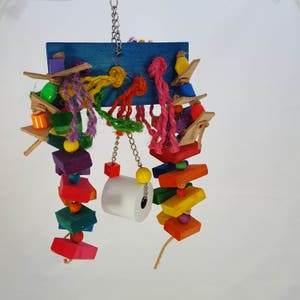 Op de afbeelding: Een kleurrijk vogelspeelgoed met een blauwe houten basis, een verscheidenheid aan houten blokken, kralen en touwen, en een witte papieren rol die aan een ketting hangt.