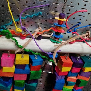 May include: A colorful wooden bird toy with a variety of textures and shapes. The toy features a wooden dowel with hanging wooden blocks, a bell, and a spinning wheel with colorful beads. The toy is designed for birds to play with and provides enrichment for their environment.