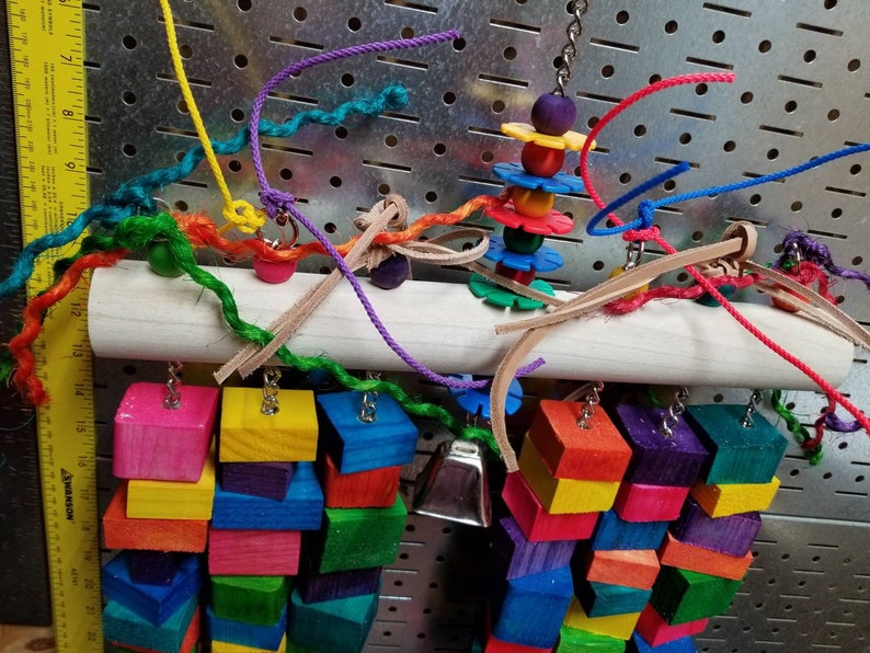 May include: A colorful wooden bird toy with a variety of textures and shapes. The toy features a wooden dowel with hanging wooden blocks, a bell, and a spinning wheel with colorful beads. The toy is designed for birds to play with and provides enrichment for their environment.