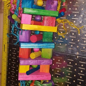 May include: A colorful wooden bird toy with a chain for hanging. The toy is made of various colored wooden blocks, some round, some square, and some rectangular. The toy also has colorful ropes and beads.