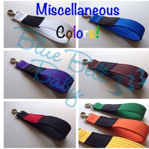 May include: A collection of colorful keychains, each featuring a textured strap in various colors: white, blue, purple, brown, black, green, orange, and yellow. Each keychain has a black accent and a silver clasp. The image includes the text "Miscellaneous Colors!"