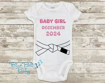 May include: White baby bodysuit with the text "BABY GIRL DECEMBER 2024" in pink. A black and white karate belt graphic is below the text. The bodysuit has short sleeves and a snap closure.