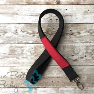 May include: Black lanyard with a red accent and a silver swivel clip. The lanyard is on a white wooden background.