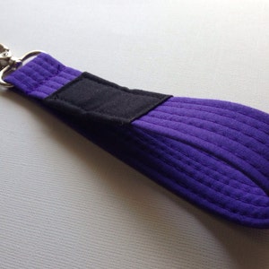 以下が含まれることがあります： 銀色の金属製の留め具が付いた、紫と黒のキーホルダー。紫色の生地はキルティングされており、黒い生地は滑らかです。