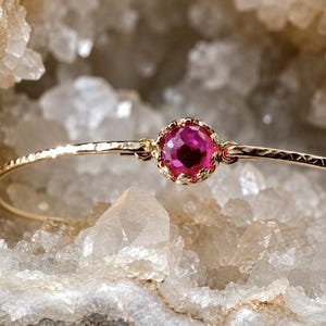 May include: A delicate gold bangle bracelet featuring a single round, faceted, vibrant pink gemstone set in a scalloped gold bezel. The thin metal band has a textured, hammered finish that catches the light, creating a refined and elegant appearance.