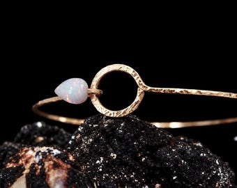 May include: A gold-toned bangle bracelet featuring a hammered texture on the thin metal band. The bracelet includes a circular centerpiece and a single teardrop-shaped white opal gemstone with iridescent flashes of color attached to the side.