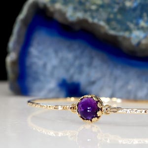 May include: A thin gold bangle bracelet featuring a single round, deep purple amethyst gemstone set in a textured, scalloped gold bezel. The band of the bracelet has a hammered, reflective finish that catches the light along its circumference.