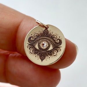 May include: Gold charm with an intricate eye design and a small clear stone in the center. The eye is surrounded by a decorative floral pattern.