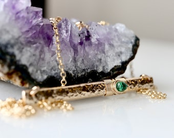 May include: A gold bar necklace featuring a hammered texture and a single small round green gemstone set in the center. The bar is wrapped with thin gold wire and suspended from a delicate gold chain, resting against a large purple amethyst cluster.
