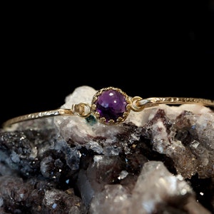 May include: A gold-toned bracelet featuring a single, round, faceted purple gemstone set in a scalloped bezel. The band has a textured, hammered finish and connects to the central stone setting with small circular loops on either side.