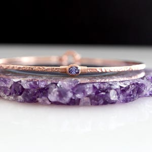 May include: A rose gold bangle bracelet with a hammered texture and a central oval setting containing a small, faceted purple stone. The bracelet features a wire-wrapped detail on either side of the stone and a small circular clasp mechanism.