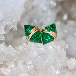 May include: A pair of stud earrings featuring two faceted, emerald-green, pointed crystals. Each crystal is wrapped with thin, metallic, rose-gold-colored wire that secures the stone and forms the base of the earring design.