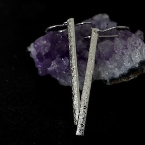 May include: A pair of silver earrings with a textured, hammered finish. The earrings are long and slender, and they are hanging from a cluster of amethyst crystals.