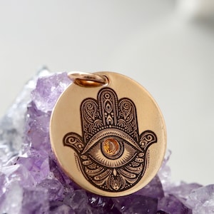 May include: A circular, gold-toned pendant with an engraved Hamsa hand design and an eye detail. It has a small loop for a chain. The background features a cluster of purple crystals. The pendant is likely for jewellery.