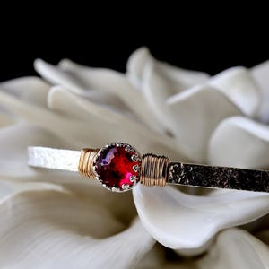 May include: A silver cuff bracelet with a hammered texture, featuring a round, faceted deep red gemstone set in a scalloped bezel. The stone is flanked by decorative gold-colored wire wrapping on both sides of the central setting.