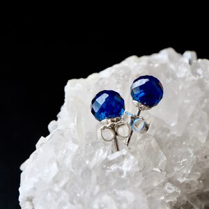 May include: A pair of stud earrings featuring round, faceted, deep blue stones set in silver-toned metal. The earrings have standard post backs and are positioned resting on a cluster of translucent, jagged white quartz crystal formations.