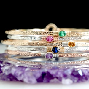 May include: A stack of five hammered metal bangles in gold, silver, and rose gold tones. Each bangle features a single small oval gemstone in green, pink, yellow, blue, or purple, secured by thin wire wrapping at the center of the band.