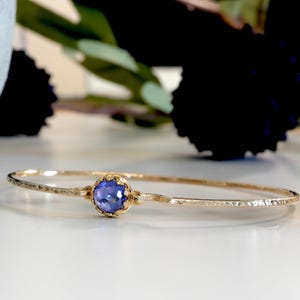 May include: A delicate gold bangle bracelet with a textured surface. The bracelet features a round, bezel-set, blue gemstone at its center. The background includes blurred greenery and a decorative object.