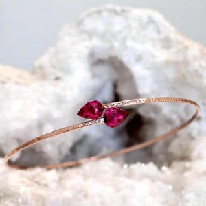 May include: A thin, rose gold-toned bangle bracelet featuring a hammered, textured finish. The open-ended design is accented by two vibrant, pear-shaped, faceted magenta-red stones positioned at the centre, creating a delicate and refined look.