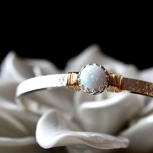 May include: A silver cuff bracelet featuring a central round white opal stone set in a scalloped crown setting. The stone is flanked by gold wire wrapping on both sides, and the silver band has a textured, hammered finish throughout its length.