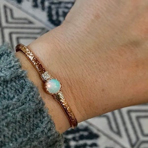 May include: A rose gold cuff bracelet featuring a textured, hammered metal finish and a single round, iridescent opal gemstone set in a prong setting. The bracelet is worn on the wrist, showcasing the reflective quality of the stone and metal.