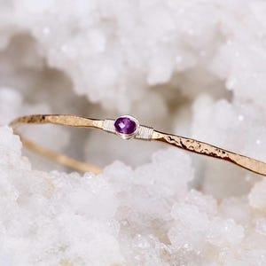 May include: A thin, hammered gold-tone metal bangle bracelet featuring a small, oval-shaped, faceted purple gemstone at the centre. The gemstone is secured by delicate silver-tone wire wrapping on both sides of the stone along the band.
