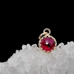 May include: A small, round pendant featuring a faceted, deep pink gemstone set in a gold-toned metal frame with a scalloped edge. The pendant includes a small circular jump ring at the top for attachment, resting on a cluster of clear crystals.