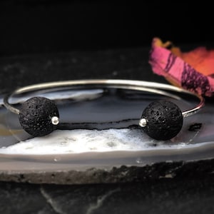 May include: A silver-toned open cuff bracelet featuring two spherical black lava stone beads at each end. The metal band is thin and polished, while the porous, textured lava beads are secured with small silver-toned metal caps on the inner sides.