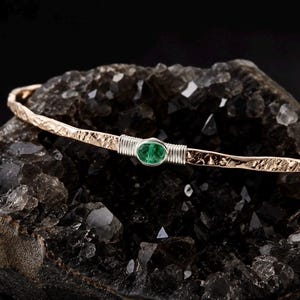 Puede incluir: Un brazalete de oro rosa martillado con una piedra preciosa verde de forma ovalada engastada en un bisel de tono plateado. La banda de metal tiene una superficie texturizada e irregular y está asegurada a ambos lados con alambre de plata.