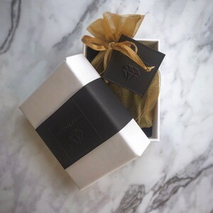 May include: A white gift box with a black label that says "remarkable" and a diamond logo. The box is open and a small gold pouch with a black tag that says "remarkable" and a diamond logo is inside.