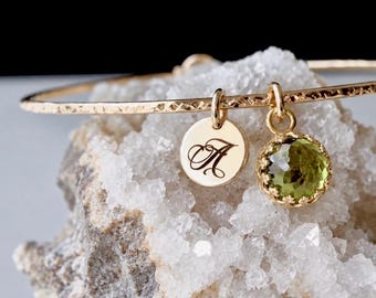 May include: A gold-toned bangle bracelet with a hammered texture, featuring two dangling charms: a small circular gold disc engraved with the letter A in a script font and a round, faceted, light green gemstone set in a gold-toned bezel.