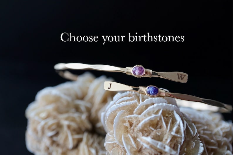 May include: Two gold bracelets with birthstones and engraved initials. The top bracelet has a purple birthstone and the letter W engraved on it. The bottom bracelet has a blue birthstone and the letter J engraved on it. The bracelets are on a white, textured surface. Choose your birthstones.