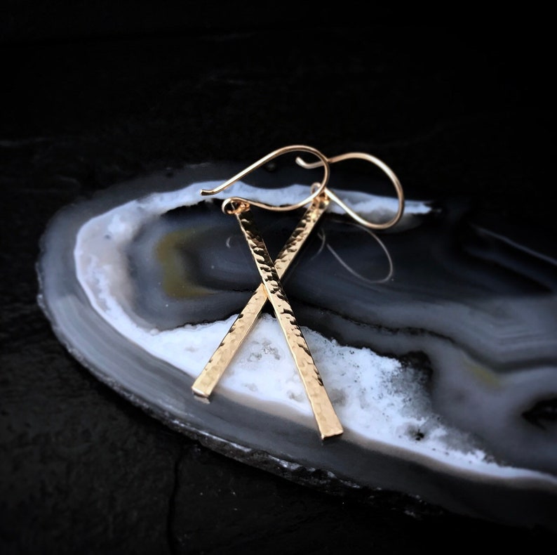 May include: Gold hammered bar earrings with a simple wire hook closure. The earrings are long and dangle.