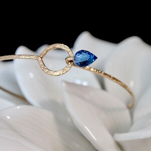 May include: A gold-toned bangle bracelet featuring a hammered texture, a circular open link, and a single pear-shaped blue gemstone accent. The metal band has a delicate, thin profile that curves to form a loop, showcasing a faceted blue stone.