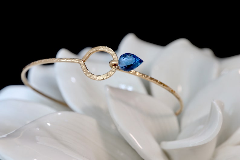 May include: A gold-toned bangle bracelet featuring a hammered texture, a circular open link, and a single pear-shaped blue gemstone accent. The metal band has a delicate, thin profile that curves to form a loop, showcasing a faceted blue stone.