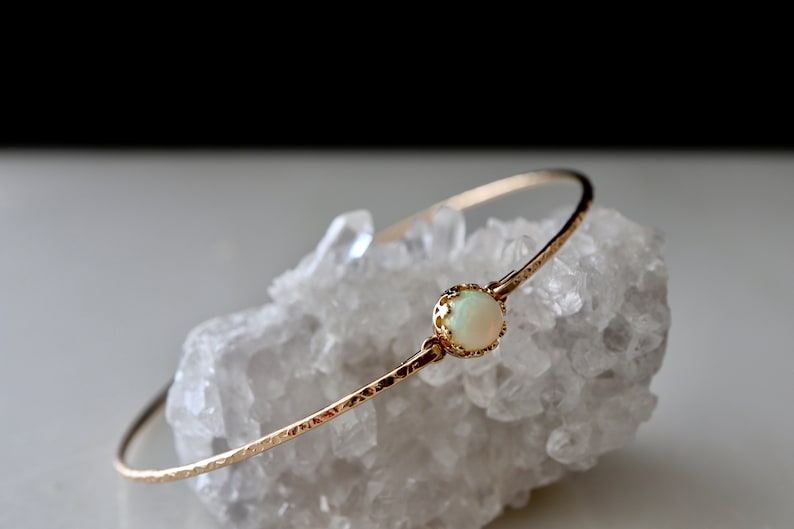 Puede incluir: Un delicado brazalete de oro con textura martillada. El brazalete presenta una piedra de &oacute;palo redonda, blanquecina, engastada en un bisel decorativo. La joya se exhibe sobre una superficie cristalina blanca.