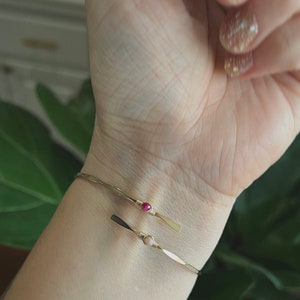 May include: Two gold bracelets with small pink and red gemstones on a person's wrist.