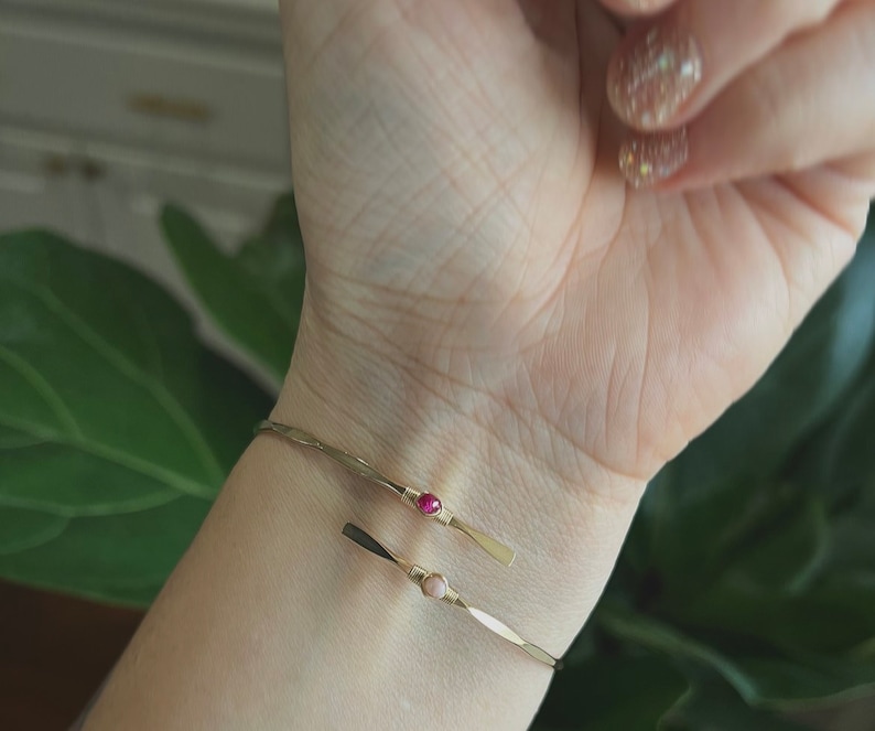 May include: Two gold bracelets with small pink and red gemstones on a person's wrist.