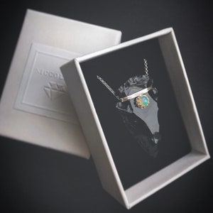 Op de afbeelding: Een zilveren ketting met een delicate ketting en een hanger met een groene opaal-steen die in een gouden bezel is gezet. De hanger is gevormd als een kleine pijlpunt en ligt in een zwarte fluwelen doos.