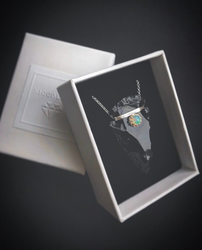 Op de afbeelding: Een zilveren ketting met een delicate ketting en een hanger met een groene opaal-steen die in een gouden bezel is gezet. De hanger is gevormd als een kleine pijlpunt en ligt in een zwarte fluwelen doos.