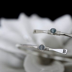 May include: Two silver wire wrapped bracelets with blue stones and initial charms. The top bracelet has the letter "A" and the bottom bracelet has the letter "B".