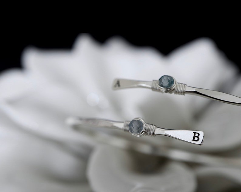 May include: Two silver wire wrapped bracelets with blue stones and initial charms. The top bracelet has the letter "A" and the bottom bracelet has the letter "B".