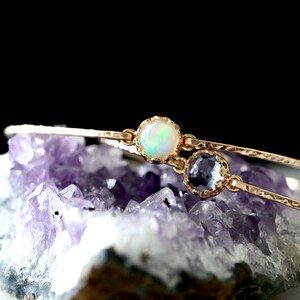 Pode incluir: Um delicado bracelete de ouro com duas pedras preciosas. O bracelete est&aacute; sobre um leito de cristais de ametista roxa. Uma das pedras &eacute; uma opala branca, a outra &eacute; uma pedra azul claro.
