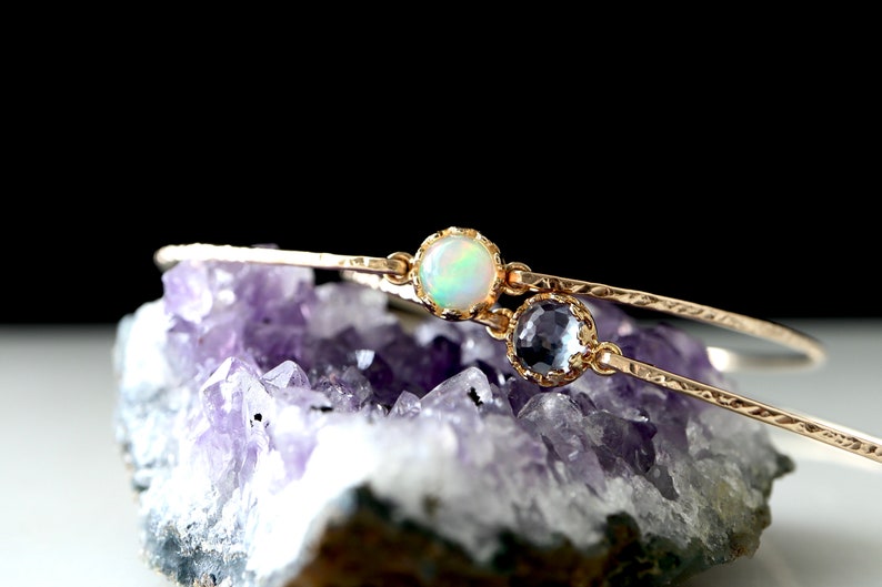 Pode incluir: Um delicado bracelete de ouro com duas pedras preciosas. O bracelete est&aacute; sobre um leito de cristais de ametista roxa. Uma das pedras &eacute; uma opala branca, a outra &eacute; uma pedra azul claro.