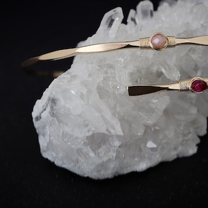 May include: Two gold wire wrapped bracelets with a pink and a red gemstone. The bracelets are displayed on a white crystal cluster.