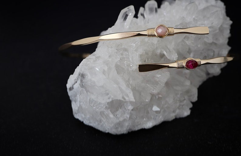 May include: Two gold wire wrapped bracelets with a pink and a red gemstone. The bracelets are displayed on a white crystal cluster.