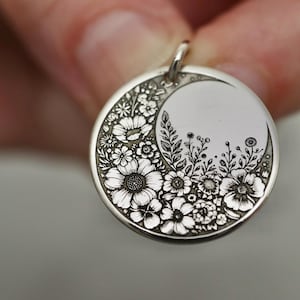 May include: A circular silver pendant featuring an intricate engraved design of various flowers and foliage arranged in a crescent moon shape. The metal surface has a polished finish with darkened recessed areas that highlight the floral details.