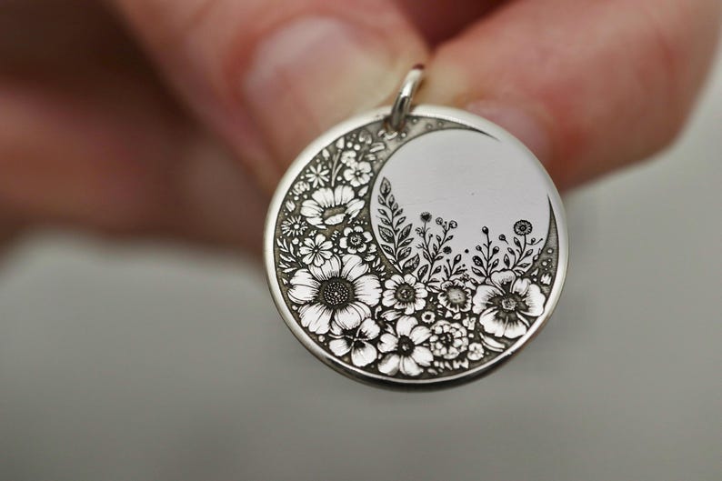 May include: A circular silver pendant featuring an intricate engraved design of various flowers and foliage arranged in a crescent moon shape. The metal surface has a polished finish with darkened recessed areas that highlight the floral details.