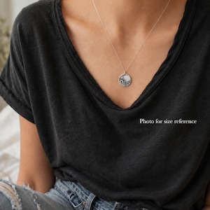 May include: A silver necklace featuring a small, round pendant with a detailed floral engraving. The pendant hangs from a delicate, thin silver chain, resting against a dark charcoal gray v-neck t-shirt and blue denim jeans.